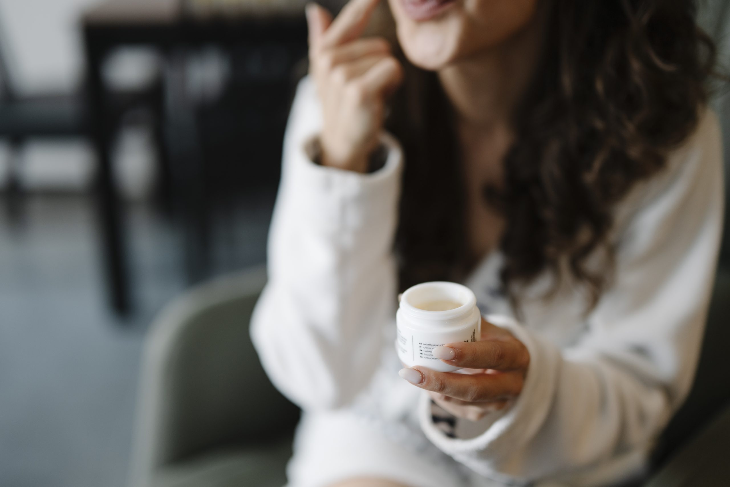 A woman applying cream to her cheek while holding an open skincare jar, showing the customer experience of using a cosmetic product.