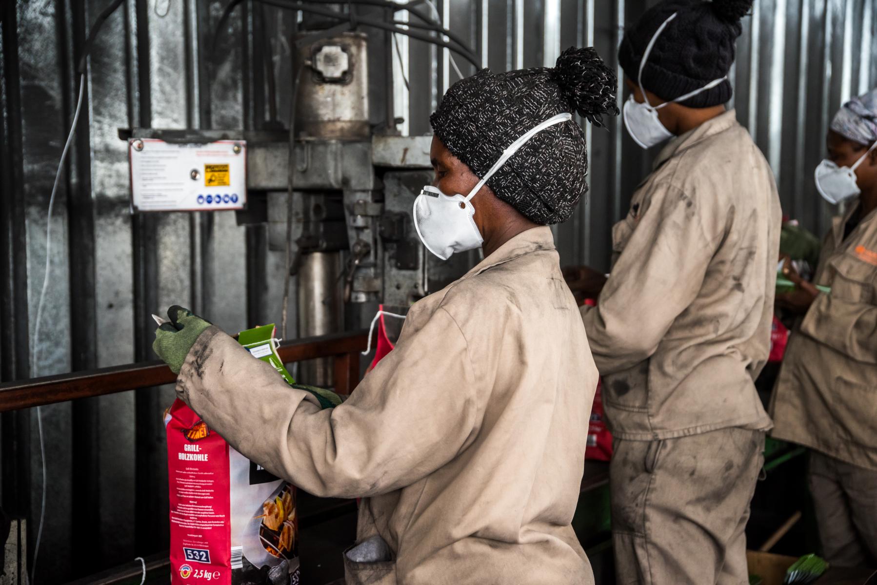Three workers wearing protective masks and work clothes package charcoal bags along a shared production line.